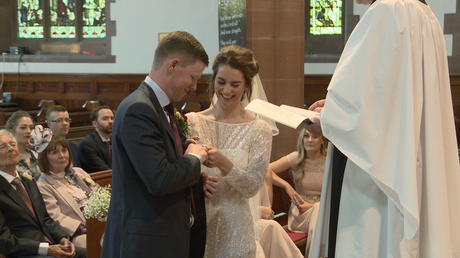 the bride and groom laugh as they try to fit the wedding rings on in front of the wedding video at their church ceremony in liverpool