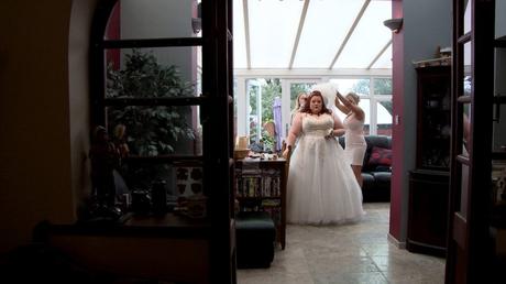 A bride stands in the conservatory of her parents house having the finishing touches added to her wedding day look with her veil being held up by the hairdresser
