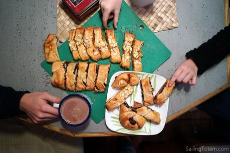 Two hands reach towards a Danish pastry while a third holds a mug of cocoa