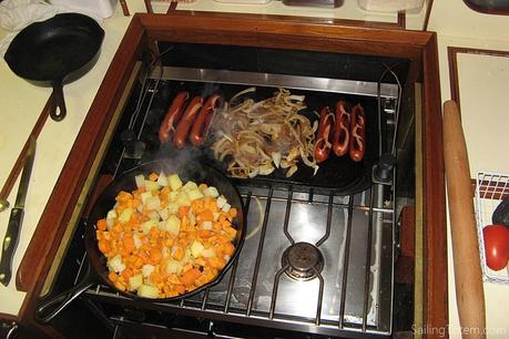 Stove with a skillet of root vegetables on the front burner and a griddle with sausages and onions  across the back