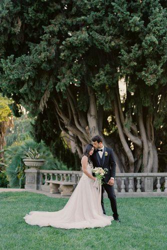 besame wedding styled shoot a bride in a white dress with a train and a bouquet of the groom in a black suit among tall trees carrie king photographer