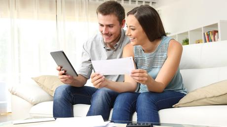 Happy couple checking bank account on line in a tablet sitting on a couch in the living room at home