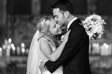 Bride smiles as groom kisses her on the cheek at The Asylum with candles in the background. 