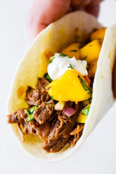 a hand holds a jerk beef taco with mango salsa on top