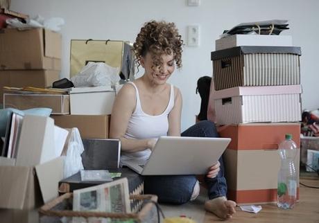 Girl typing on a laptop in a messy room full of boxes.