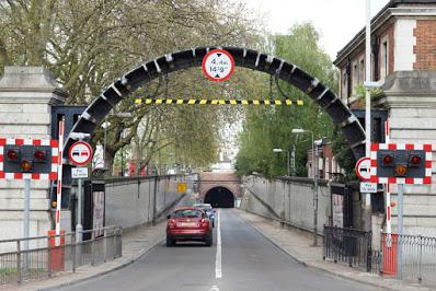 Photograph of a tunnel entrance. In the foreground is a metal arch between stone posts, with a height restriction sign on the top and a black-and-yellow horizontal bar. There are warning lights to either side, and a raised barrier. Round traffic signs indicate no overtaking for 1.25 miles. Beyond the arch are two cars driving along a narrow two-lane road towards the arched mouth of a tunnel whose interior appears dark. The approach is lined with trees and a brick building.
