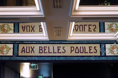 Section of a mosaic frieze saying 'Aux Belles Poules', with rose wreaths at either end of the words, and blue borders; and its reflection in a mirrored ceiling.