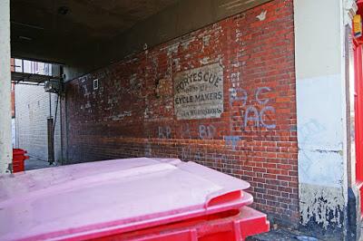 A photograph of the ghost sign and surrounding alleyway: the red brick wall, covered with graffiti tags, joins onto a flat concrete roof. To the left of the picture, a daylit yard is visible; to the right, a colourful shop front. In the foreground is the top of a large red commercial bin, bleached pink by the sun.