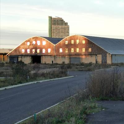 Photograph taken in late afternoon light. A single-lane private road leads to a warehouse, whose two gables are distinctively curved. Its first and second floors are of rusty corrugated iron, pierced with rows of windows. The ground floor storey is painted cream with large garage-type doors. In front of it and to the side of the road is bare soil interspersed with scrubby vegetation. Behind the warehouse, a high-rise block of flats is visible.