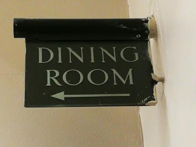 At the corner of two cream-painted walls, a round metal tube protrudes with a sign hanging from it. Both are dark brown, although marked on the right edge by cream paint left when the walls were repainted, and the sign says 'DINING ROOM' in pale letters with an arrow pointing left.