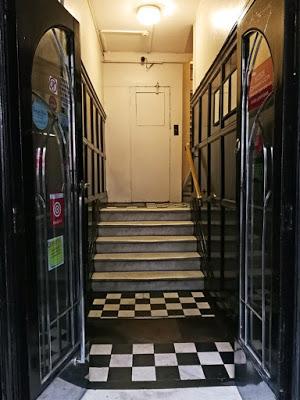 Two black doors with large glass panels are open, leading to a tiled hallway with a short flight of stairs. There is a closed, unmarked door directly ahead; the stairs continue to the right.