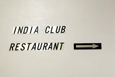 Individual black letters on pale gold backgrounds are stuck onto a cream wall. They say INDIA CLUB RESTAURANT and to their right is stuck a gold arrow on a black background,  pointing right.