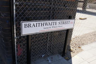 Photograph of a street sign in front of a wire fence. The modern sign is white, with black text saying 'BRAITHWAITE STREET E1 formerly Wheler Street, LONDON BOROUGH OF TOWER HAMLETS'
