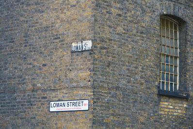 Photograph of the corner of a brick building. On one wall are two signs, both saying LOMAN STREET SE1'. The upper sign is older and smaller; it says 'Borough of Southwark' at the top, abbreviates 'street' to 'St' and writes 'SE1' as 'S.E.1'. The lower sign is a modern one. On the other wall is a tall, narrow arched window divided into many small panes.