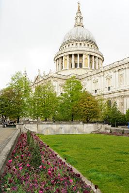 Photograph of St Paul's cathedral taken from the south-east. The south facade and famous dome are visible. In the foreground is a lawn bordered by flower beds filled with pink and purple flowers including tulips, and small, shaped conifers. The far wall of this garden has a row of bronze lion heads spouting water.