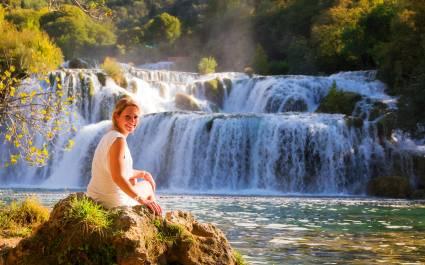 Stunning young woman sitting at a waterfall in Krka national park Wearing a white dress in summer, smiling lovely over her shoulder - Croatia and Slovenia Travel Guide