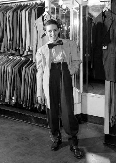 period photo of young teenage kid wearing zoot suit with wide shoulders, big bowtie, and long watch chain standing looking cool in men's clothing store