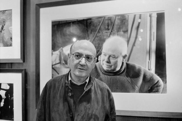 Theo ANGELOPOULOS posing in front of a portrait of Jean Renoir, photographed by Rene BURRI. 1998. by Guy Le Querrec