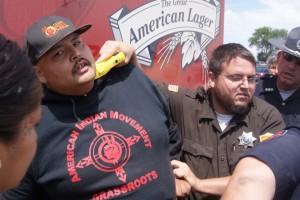 A cop holds a taser on a man’s neck as he stands behind a Budweiser truck.