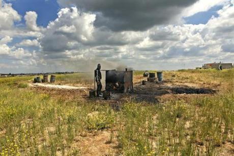 A farmhand stands nearby the home-made oil refinery, consisting of a rusted tank connected to a few tubes.