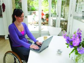 Woman sitting in wheelchair using laptop at kitchen table, smiling