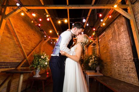 Bride and groom kiss under festoon lights