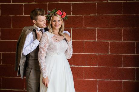 Boho style bride and groom have a cuddle in front of red brick wall at barn wedding