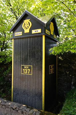 Photograph of a square, black wooden structure with yellow-painted edges, set behind a low wall with trees behind it. There are signs on it including an AA logo, 'PORLOCK HILL', and 'BOX 137'