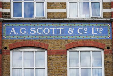 Photograph of a section of brick facade. There is a tiled sign: inside a green border is a blue background with white lettering saying 'A. G. SCOTT'. Above the sign are the bottoms of two windows and the brick is in red and white stripes. Below are the tops of two windows with curved top edges, and the wall is all red brick.