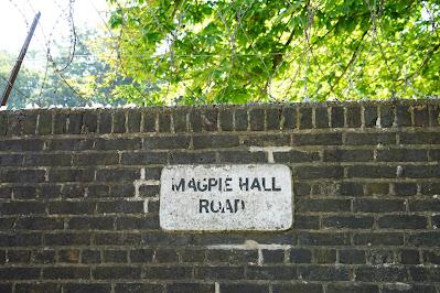 Photograph of a wall sign. It has black text reading 'MAGPIE HALL ROAD' on an off-white background. The sign is on a red brick wall topped with broken glass and loops of barbed wire.