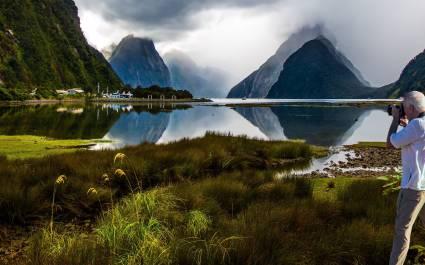 New Zealand. The famous ocean fjord Milford Sound. The gray-haired tourist photographs a picturesque landscape. Port of tourist and pleasure ships, yachts and boats.