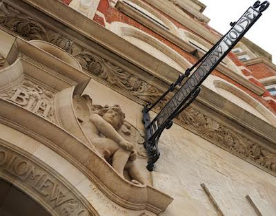 Photograph showing part of a doorway arch carved in pale golden stone, and a metal sign with the words 'Saint Bartholomew House' which extends at right angles from the facade