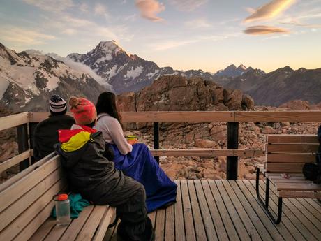 Three hikers in sleeping bags sitting on the deck of Mueller Hut with Aoraki Mt Cook in the background