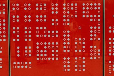 Photograph of a shiny red panel with white dots in a grid pattern. There is a square gap in the dots, with an outline of a house shape in the centre, representing Soho Square. Some of the dots are in fact Underground roundels.