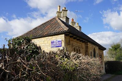 Photograph showing a small stone building with a pitched roof and gothic-arched windows. On the side wall is a blue enamel notice with the words 'Bath Humane Society's Station for Lifebuoys and Drag-Poles' visible. In the foreground is a hedge.