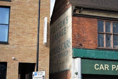 Photograph showing a recently built brick building on the left and a much older brick building on the right. There is a small gap between them, with a painted ghost sign visible on the older building's side wall.