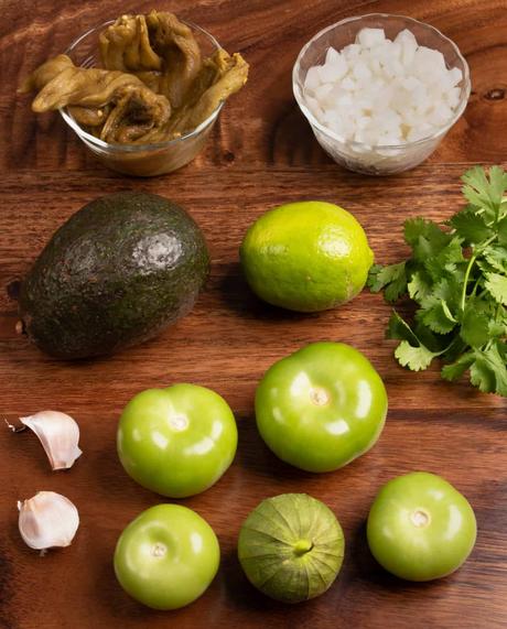 top down photo of tomatillos, avocado. Green Chiles. garlic, onion. cilantro, and a lime