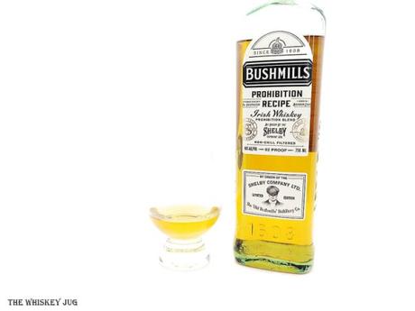 White background tasting shot with the Bushmills Prohibition Recipe Irish Whiskey bottle and a glass of whiskey next to it.