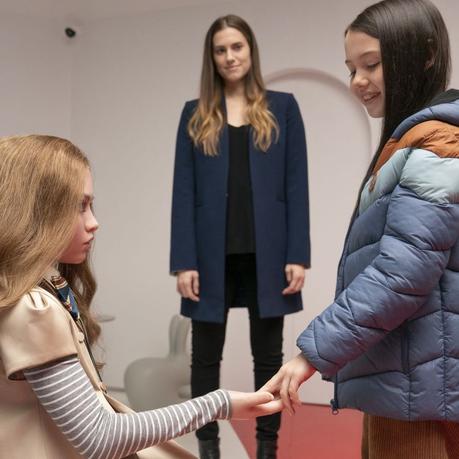 A still from M3GAN (2022) depicting Amie Donald as Megan acquainted with Violet McGraw as Cady with Allison Williams as Gemma on the background