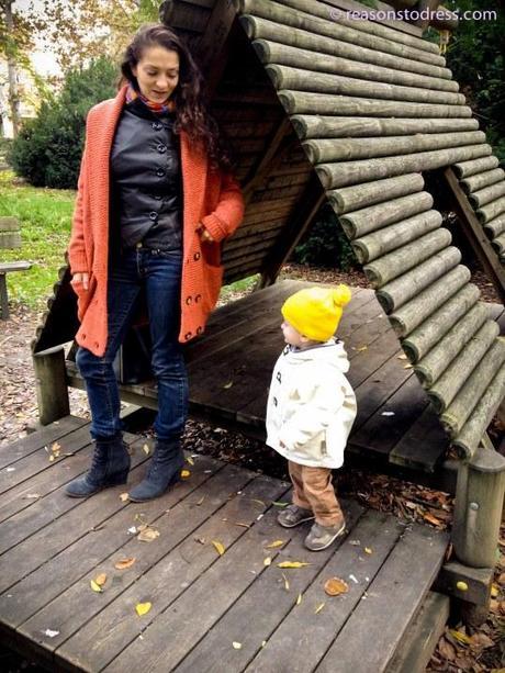 Woman wearing orange red coatigan with leather jacket leather moto jacket and jeans wedge booties blue suede shoes blue suede wedge booties toddler in yellow hat yellow pompom hat park wooden park mom and toddler mother and son mom and baby