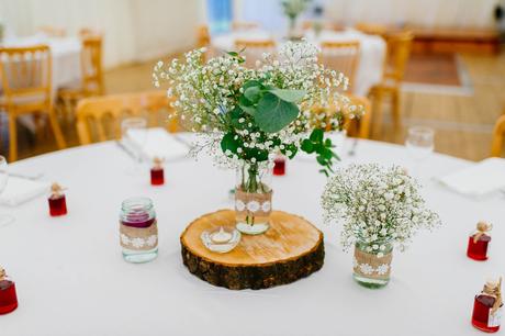 A captivating boho-inspired centrepiece on a wedding table. A rustic wood log slice serves as the base, adorned with a delicate vase filled with whimsical boho flowers. The vibrant blooms feature a mix of wildflowers, feathers, and lush greenery, creating a dreamy and enchanting display.