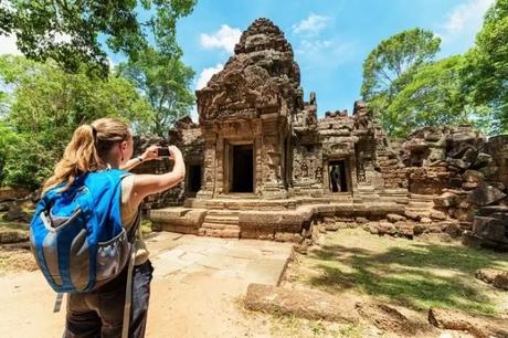 Young female tourist with smartphone taking photo of gopura under blue sky near the entrance of ancient Preah Khan temple in Angkor