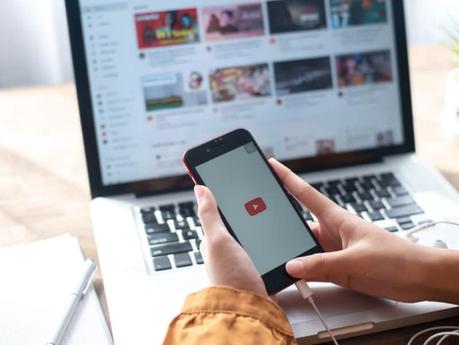 Top Tactics for Creating YouTube Engagement: An Overview! A person opens up YouTube on a phone. There is a laptop in the background.
