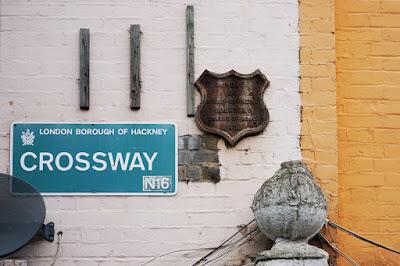 A blue sign with white text says 'LONDON BOROUGH OF HACKNEY CROSSWAYS N16'. It seems 'N16' has been placed on later. Also on the wall is a shield-shaped plaque.