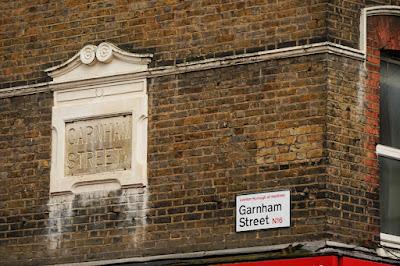 On a brick wall, a stone tablet in a carved stone surround says 'Garnham Street'. To its right is a modern sign with black and red lettering on a white backround saying 'London Borough of Hackney Garnham Street N16'; above are traces of a painted sign.