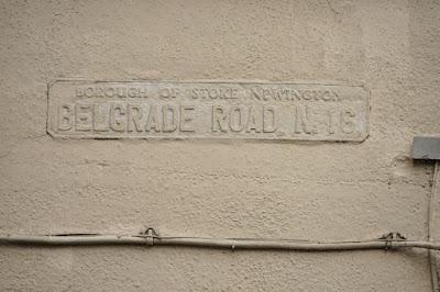 A metal sign for Belgrade Road has been painted over in the same cream paint as the wall behind, but the raised lettering is still visible.