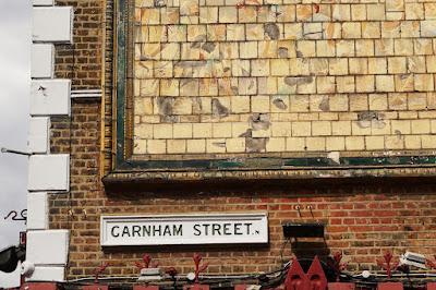 On a brick wall, a painted sign in a moulded rectangular frame says Garnham Street. Above it is part of a tiled panel with painted figures faintly visible.
