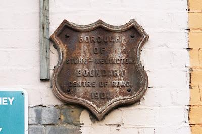 A shield-shaped metal plaque on a wall. It is streaked and darkened, but says 'Borough of Stoke-Newington boundary center of road 1901'.