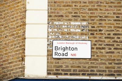A rectangular sign with black border and rounded cornders says 'London Borough of Hackney Brighton Road N16' in black and red letters. Faint painted words are visible above.