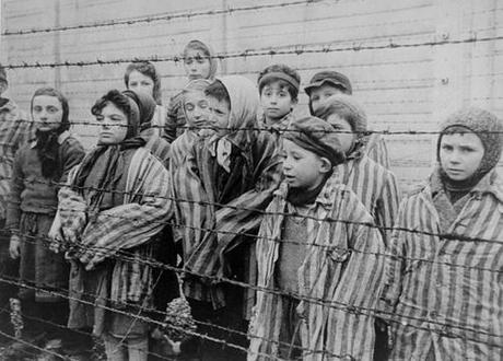  Child survivors of Auschwitz, wearing adult-size prisoner jackets, stand behind a barbed wire fence. Among those pictured are Tomasz Szwarz; Alicja Gruenbaum; Solomon Rozalin; Gita Sztrauss; Wiera Sadler; Marta Wiess; Boro Eksztein; Josef Rozenwaser; Rafael Szlezinger; Gabriel Nejman; Gugiel Appelbaum; Mark Berkowitz (a twin); Pesa Balter; Rut Muszkies (later Webber); Miriam Friedman; and twins Miriam Mozes and Eva Mozes wearing knitted hats. 1945, Auschwitz, [Upper Silesia] Poland Credit: Belarusian State Archive of Documentary Film and Photography 